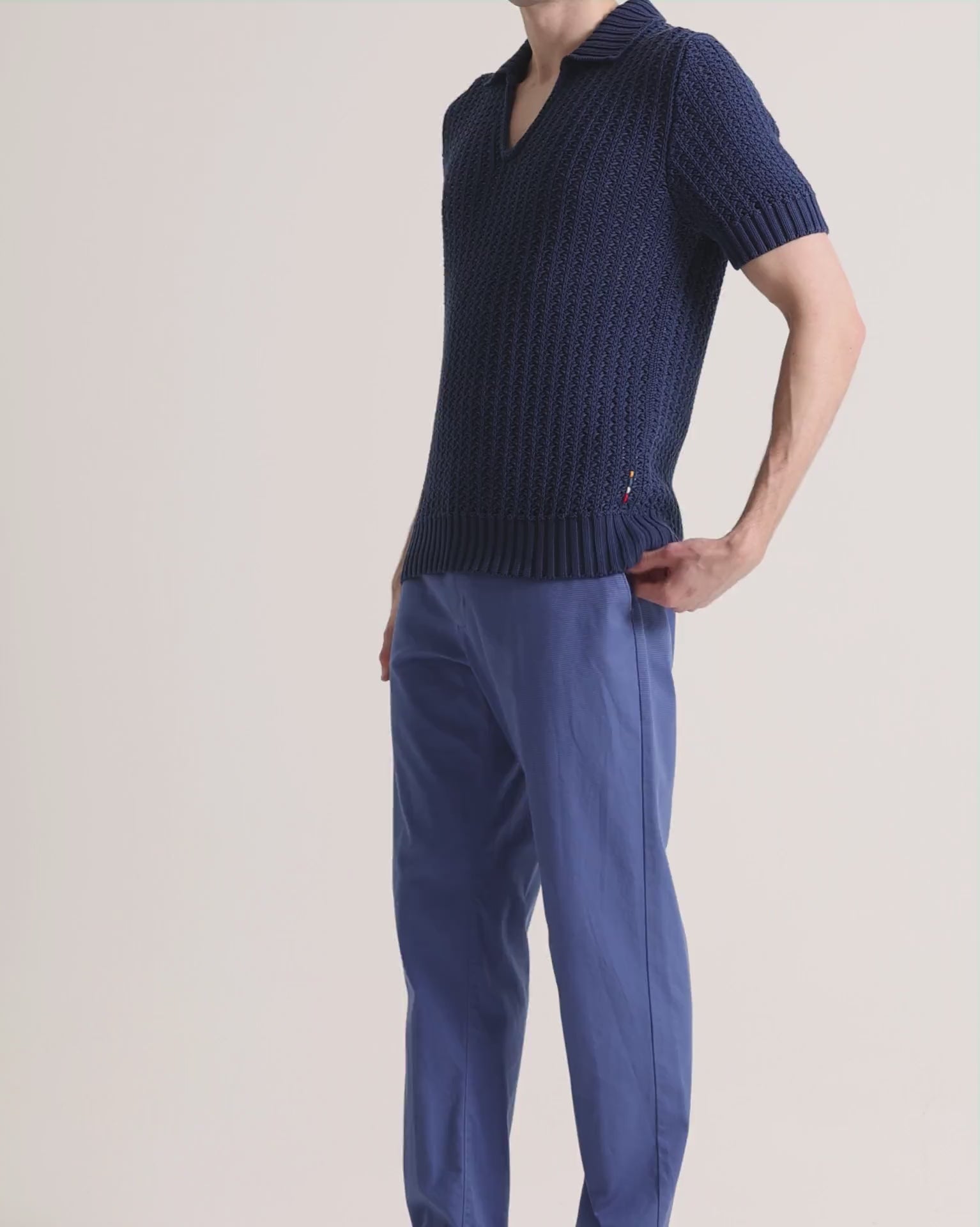 Man wearing navy knitted polo shirt with blue trousers on a plain background
