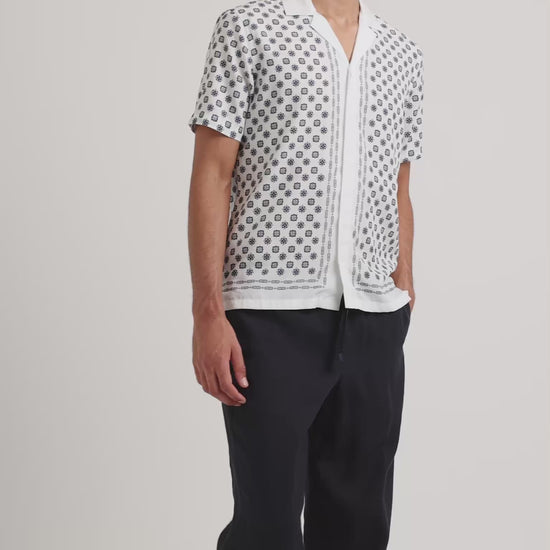 Man wearing a white geometric print shirt with black trousers on a plain background
