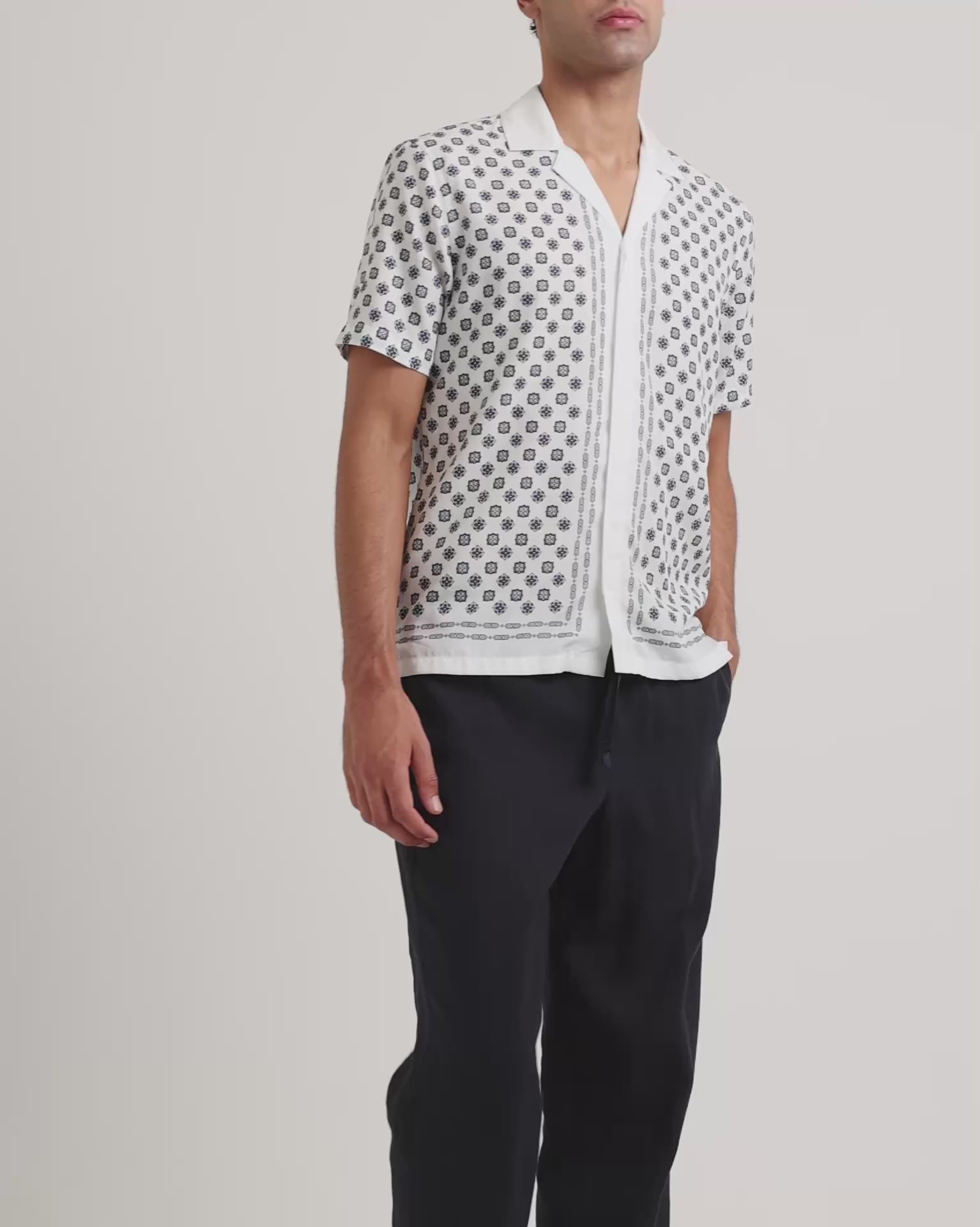 Man wearing a white geometric print shirt with black trousers on a plain background