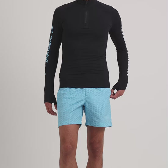 Man wearing long sleeve black rash guard with blue printed swim shorts on a plain background