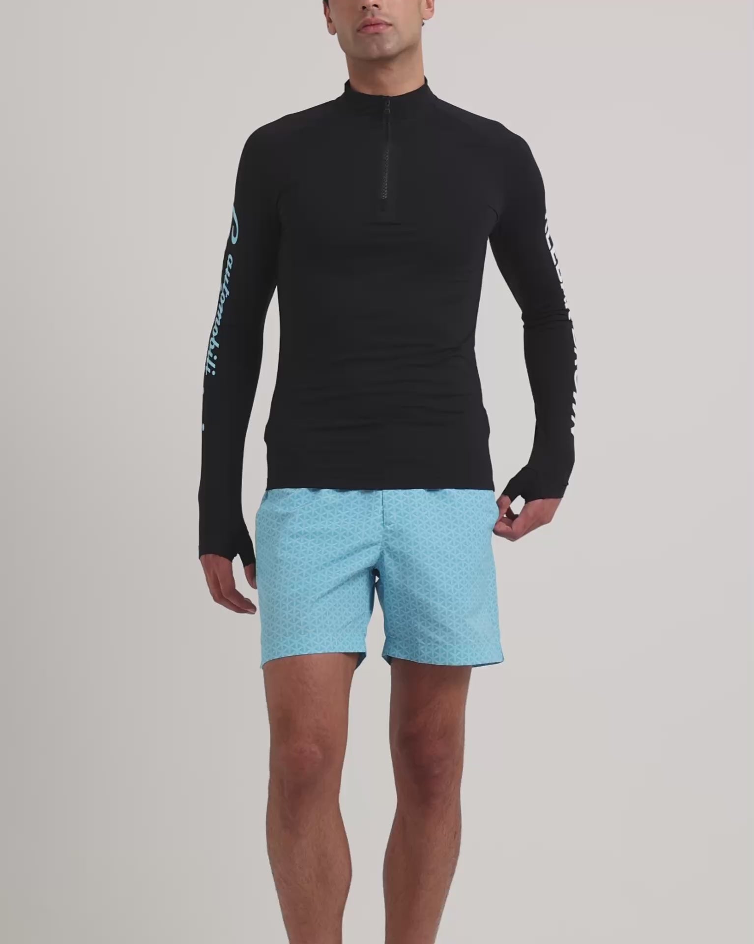 Man wearing long sleeve black rash guard with blue printed swim shorts on a plain background