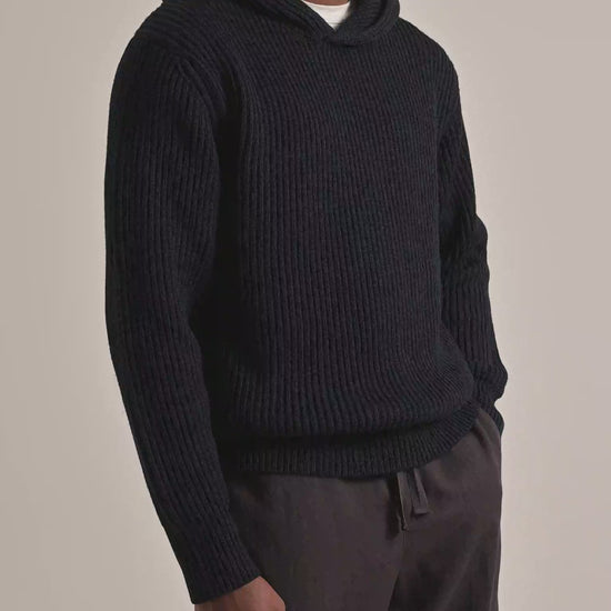 Man wearing navy knitted hoodie with brown trousers on a plain background