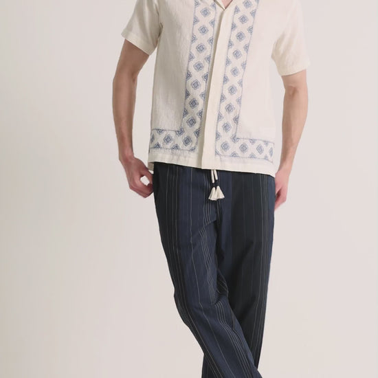 Man wearing printed white linen shirt with navy trousers on a plain background