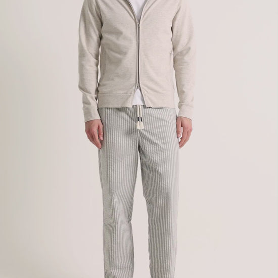 Man wearing beige zip up sweatshirt with grey striped trousers on a plain background