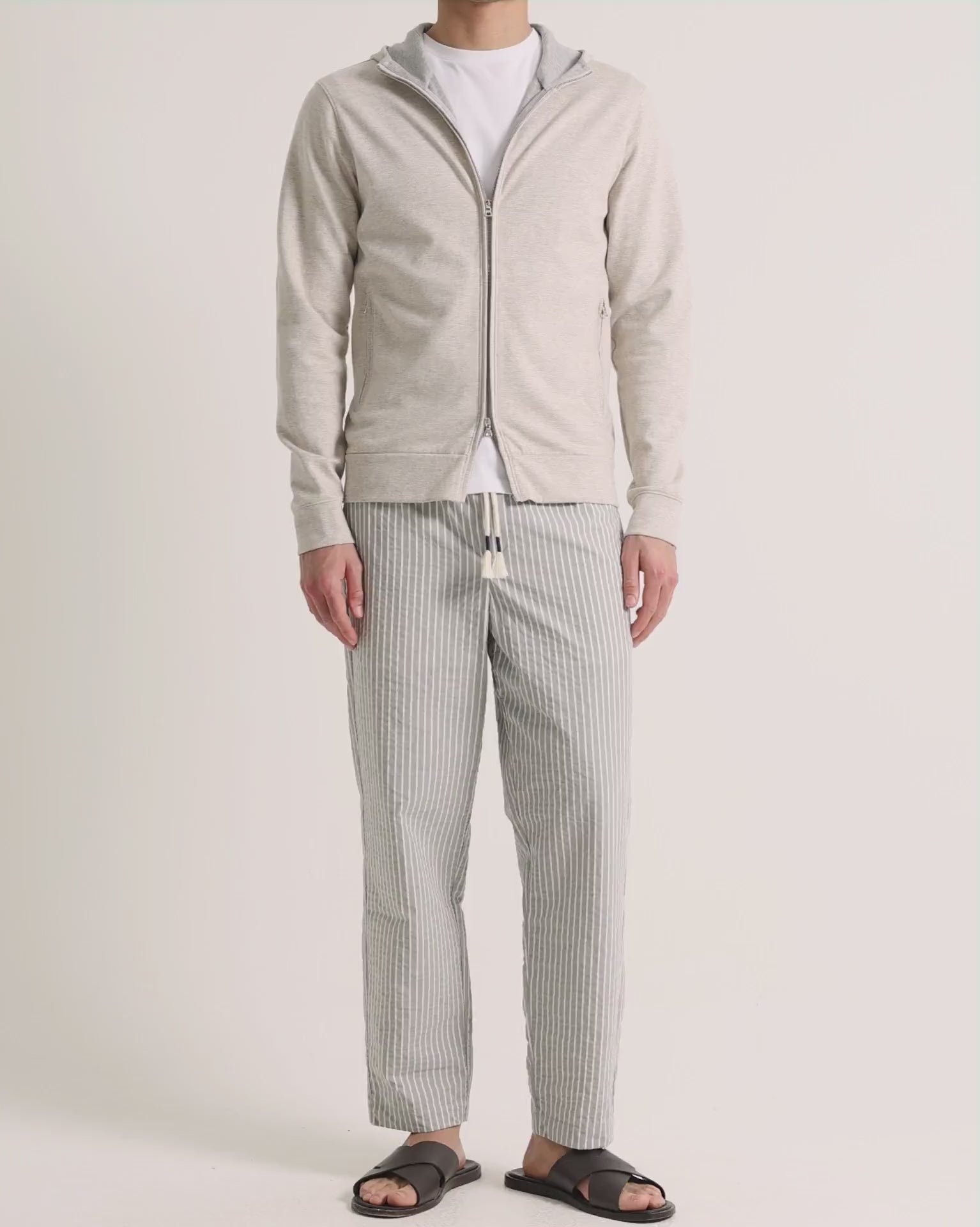 Man wearing beige zip up sweatshirt with grey striped trousers on a plain background