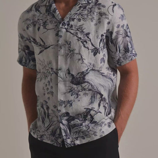 Man wearing blue botanical printed shirt with dark navy trousers on a plain background
