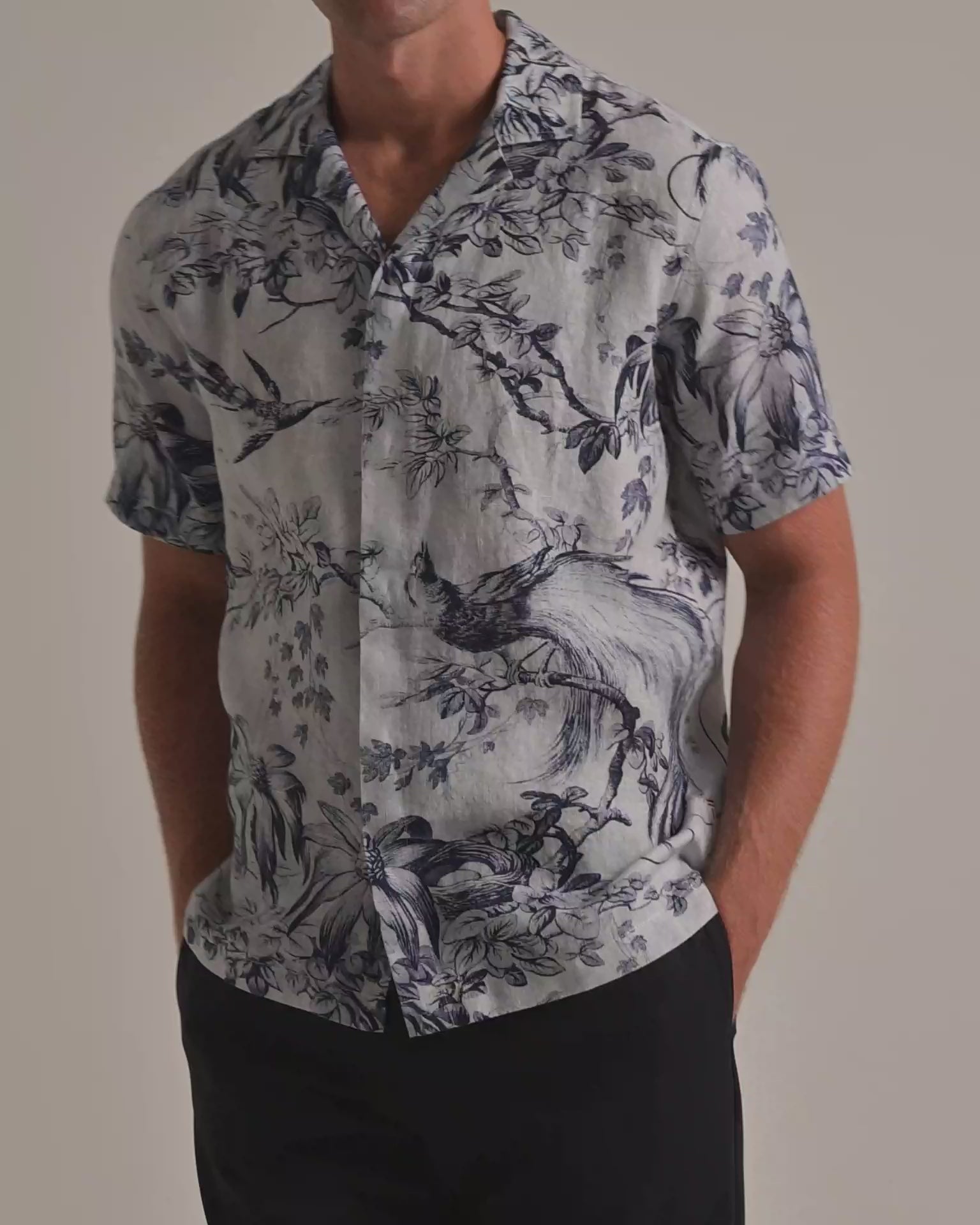 Man wearing blue botanical printed shirt with dark navy trousers on a plain background