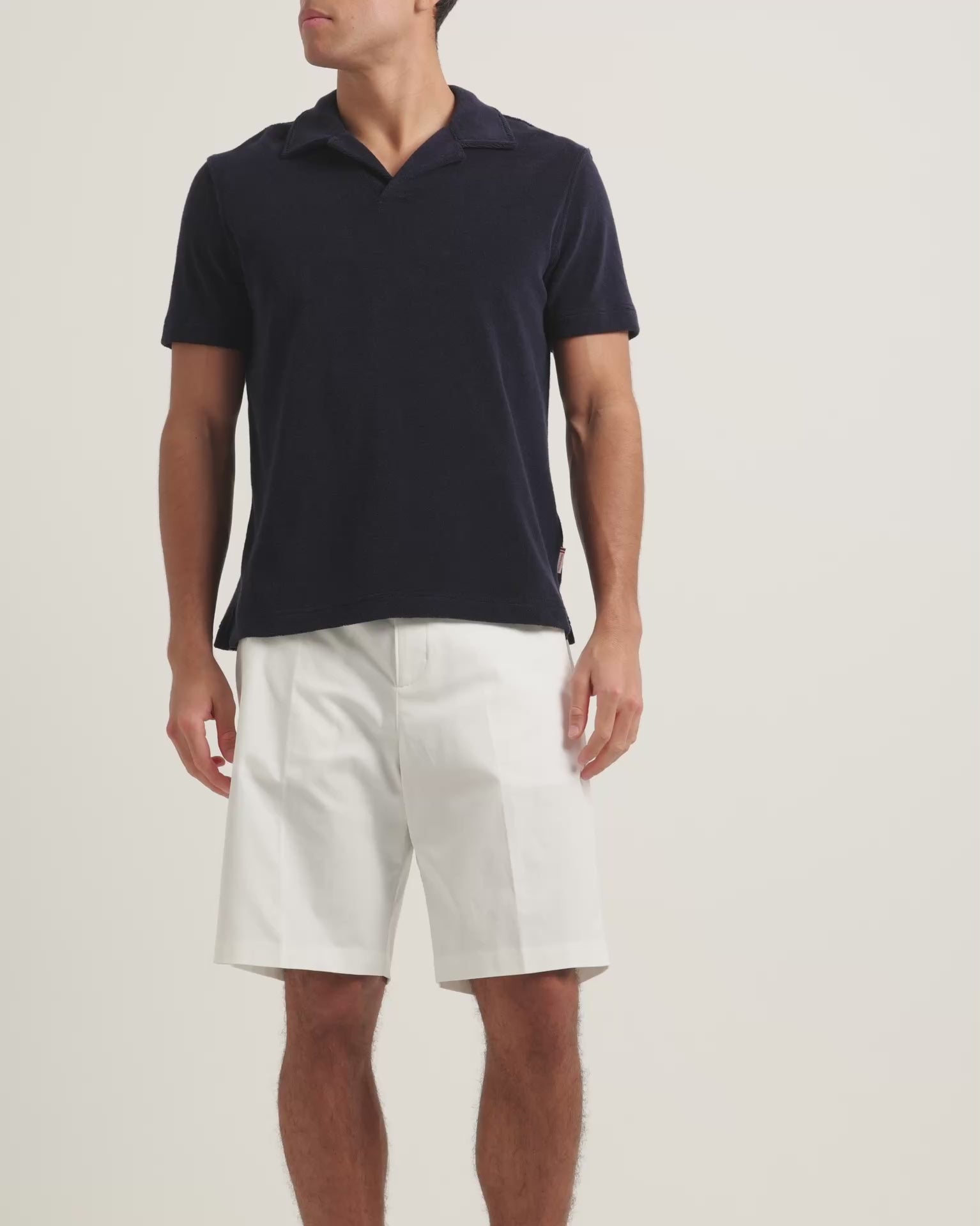 Man wearing navy towelling polo shirt with white bermuda shorts on a plain background