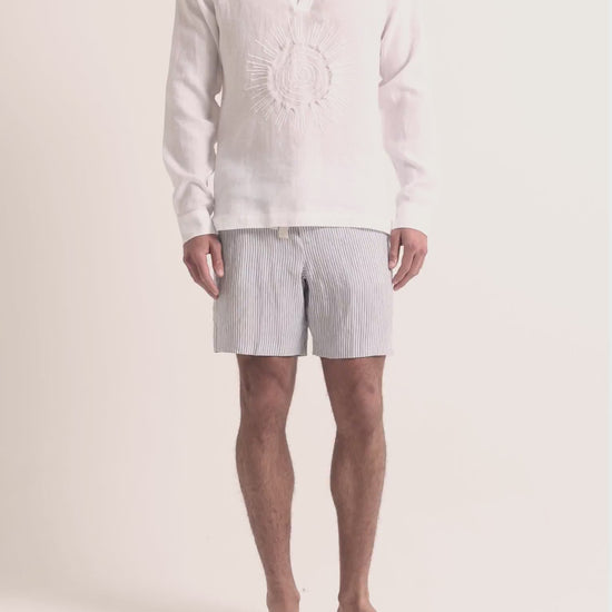 Man wearing white embroidered linen shirt with striped shorts on a plain background