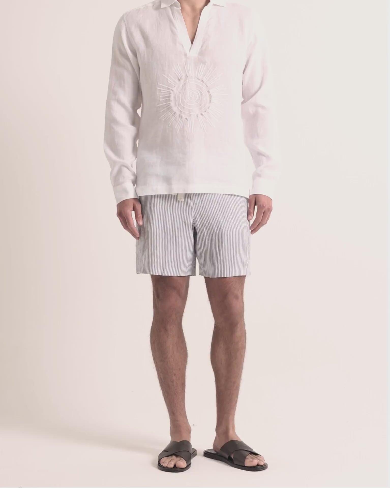 Man wearing white embroidered linen shirt with striped shorts on a plain background