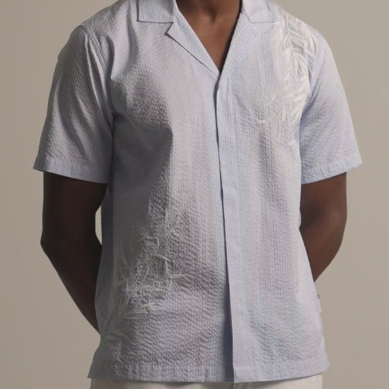 Man wearing light blue embroidered shirt with white botanical embroidery. Worn with light beige trousers against a plain background