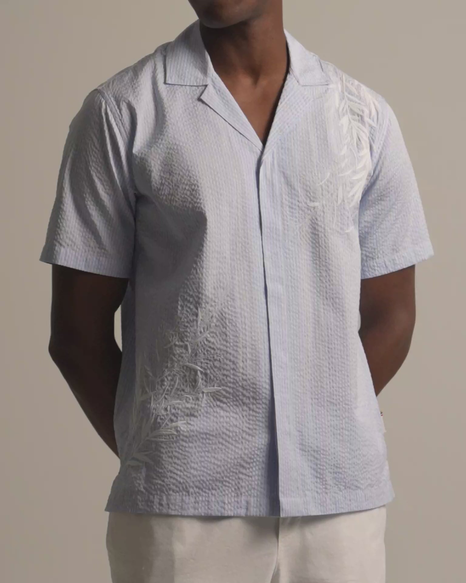 Man wearing light blue embroidered shirt with white botanical embroidery. Worn with light beige trousers against a plain background