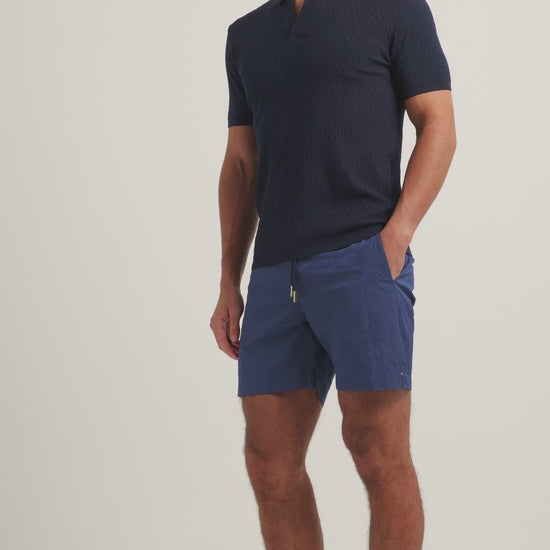 Man wearing navy textured polo shirt with navy drawcord swim shorts on a plain background