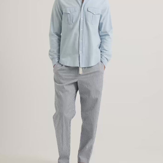 Man wearing light blue shirt with chest pockets and blue seersucker linen trousers with drawstring waist