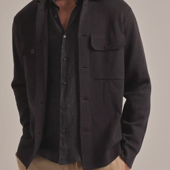 Man wearing dark brown knitted overshirt jacket with grey linen shirt and beige trousers on a plain background