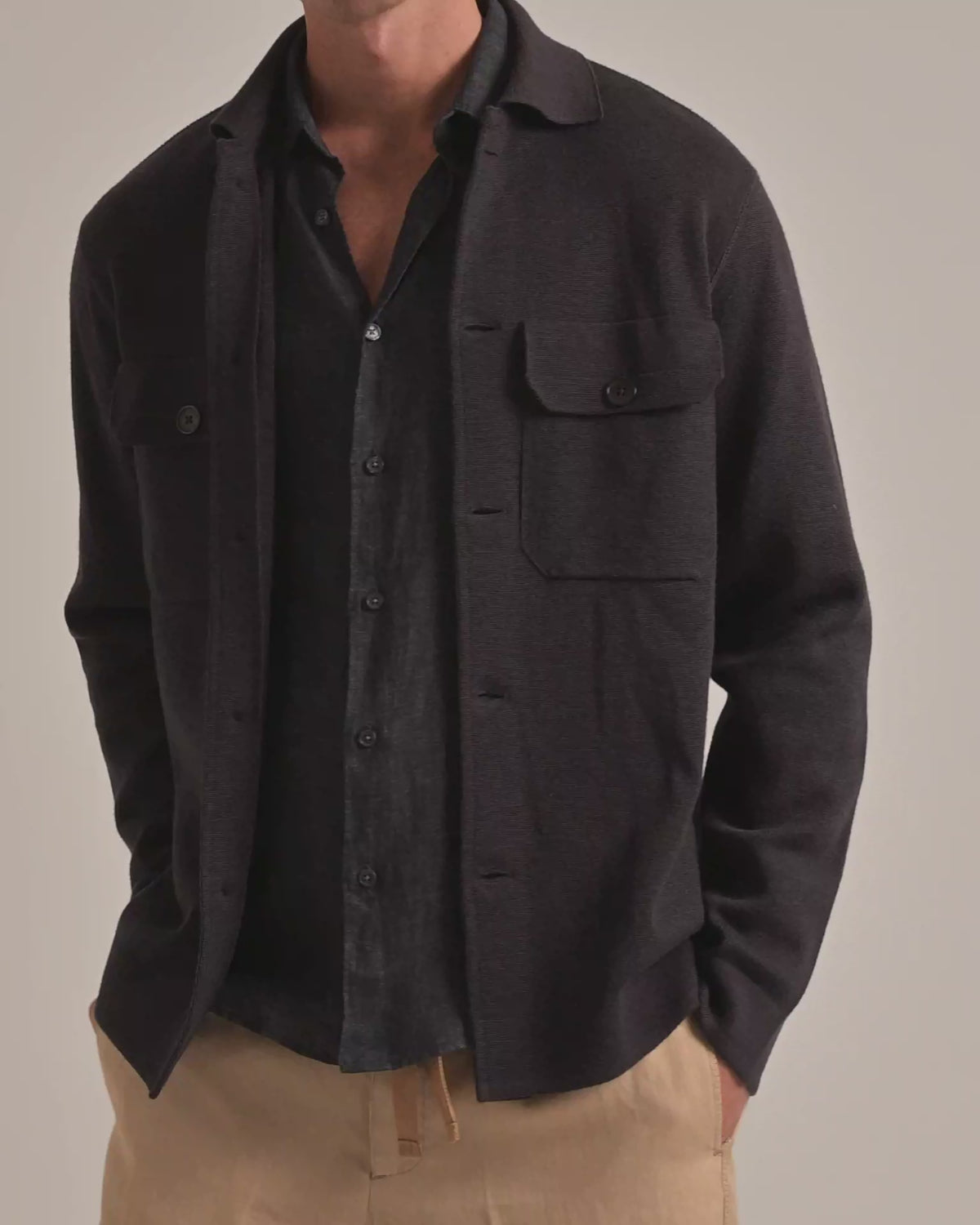 Man wearing dark brown knitted overshirt jacket with grey linen shirt and beige trousers on a plain background