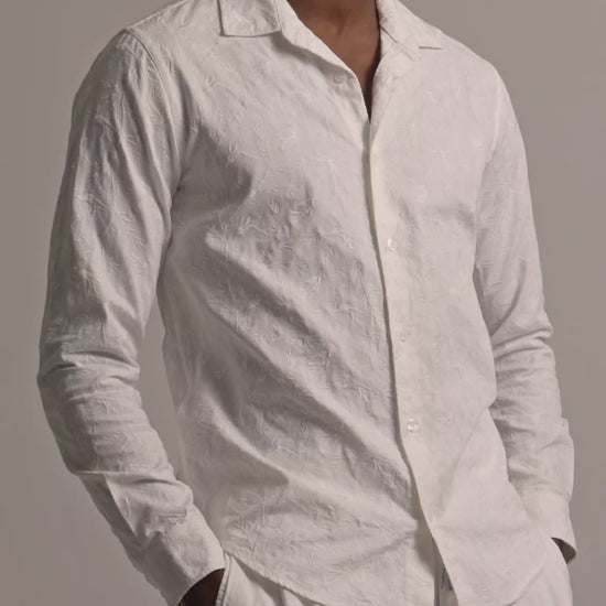 Man wearing white embroidered shirt in botanical detail with white trousers against a plain background