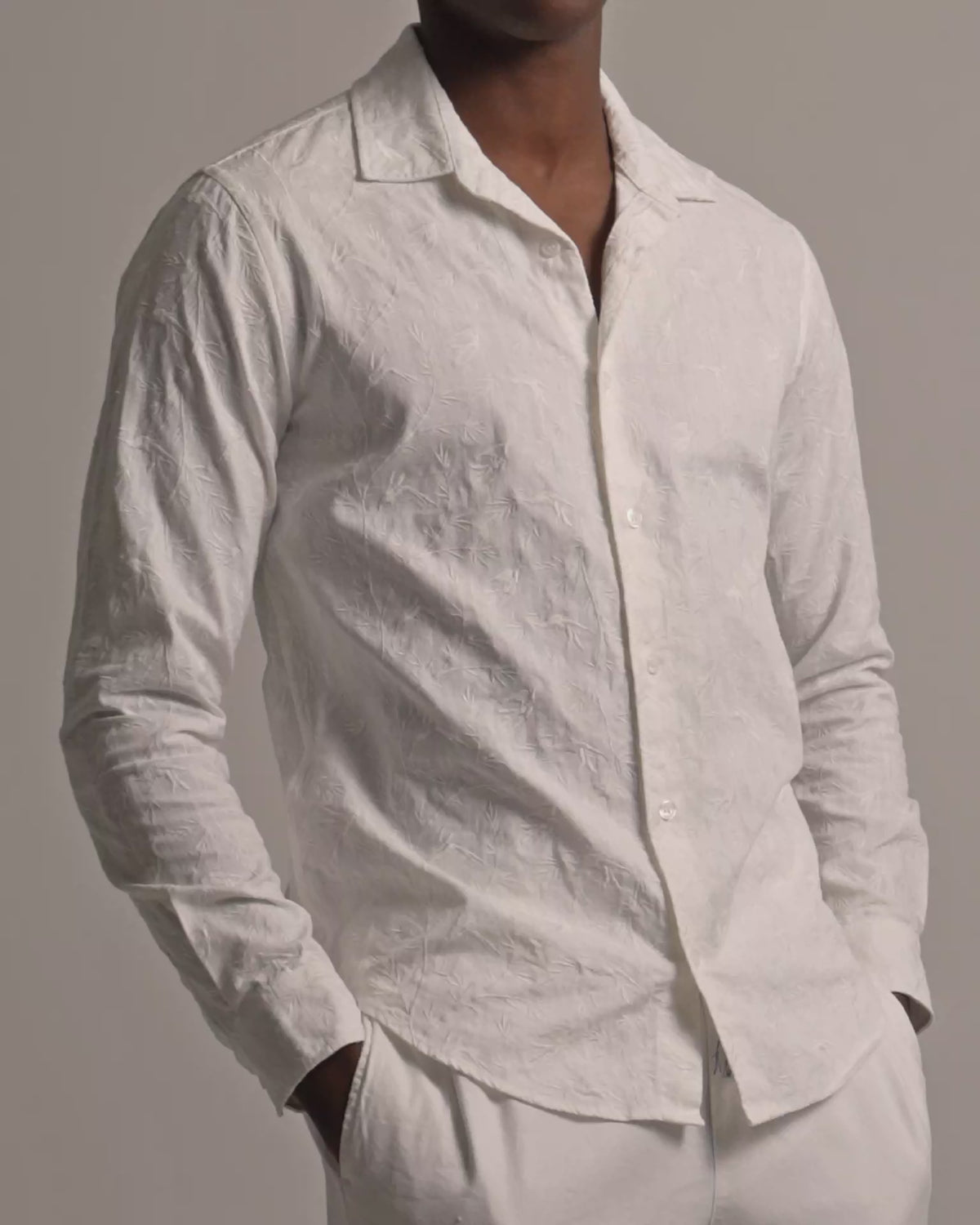 Man wearing white embroidered shirt in botanical detail with white trousers against a plain background