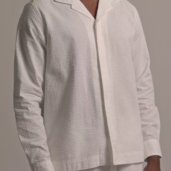 Man wearing white seersucker shirt with white trousers against a plain background