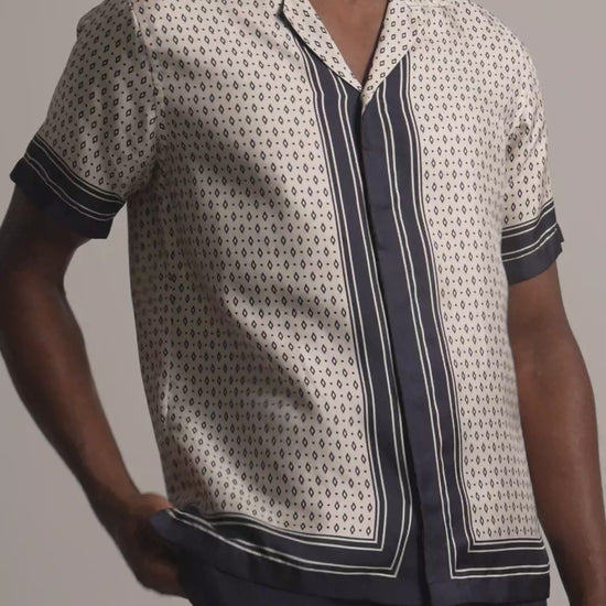 Man wearing light beige silk printed shirt with black border detail and dark trousers against a plain background