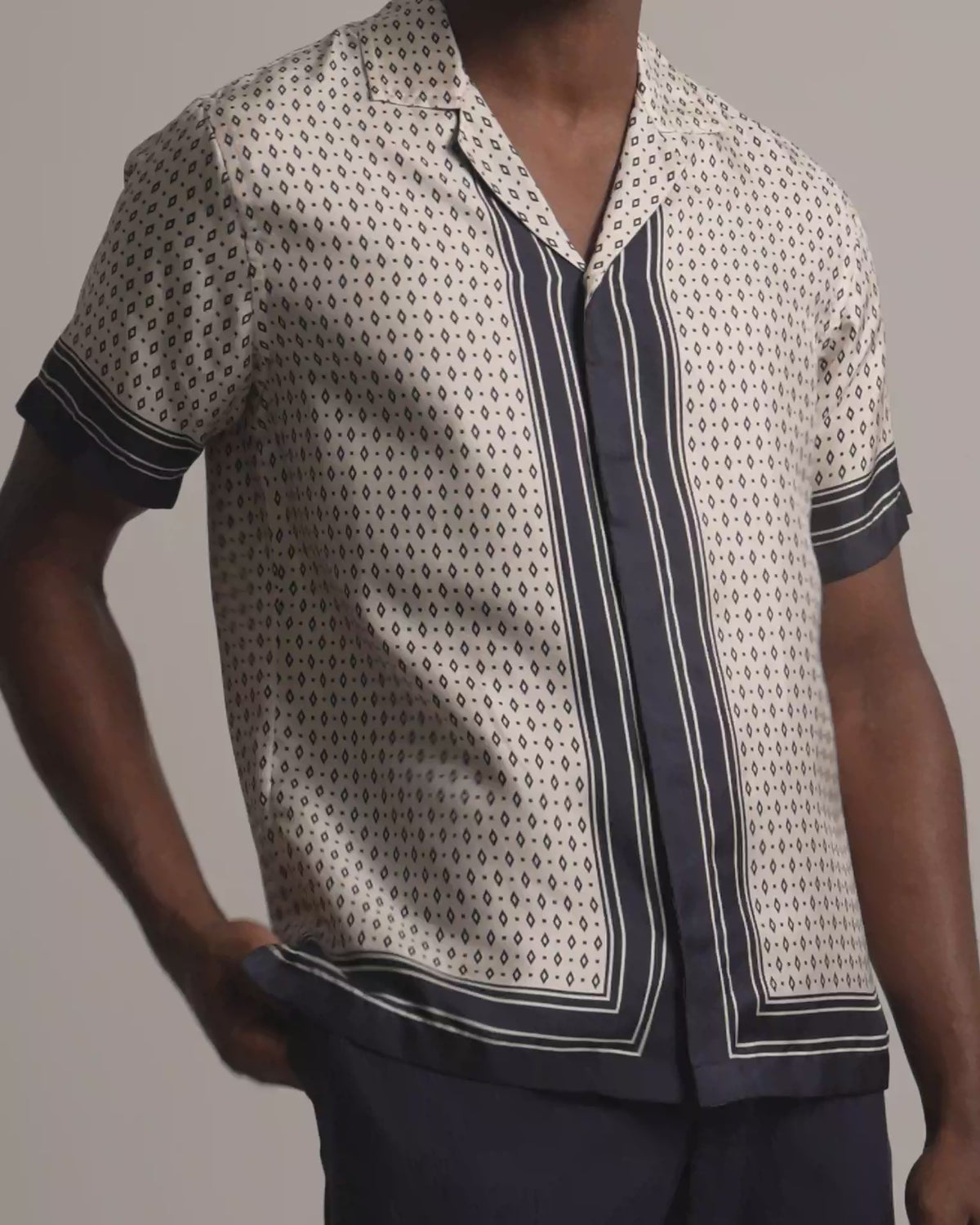 Man wearing light beige silk printed shirt with black border detail and dark trousers against a plain background