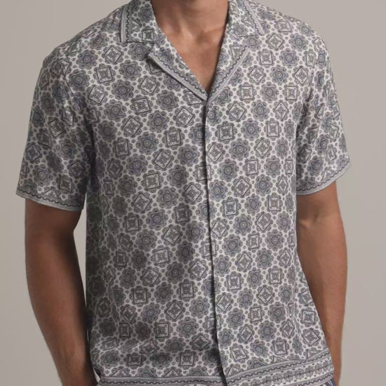 Man wearing white and grey geometric printed shirt with striped trousers against a plain background
