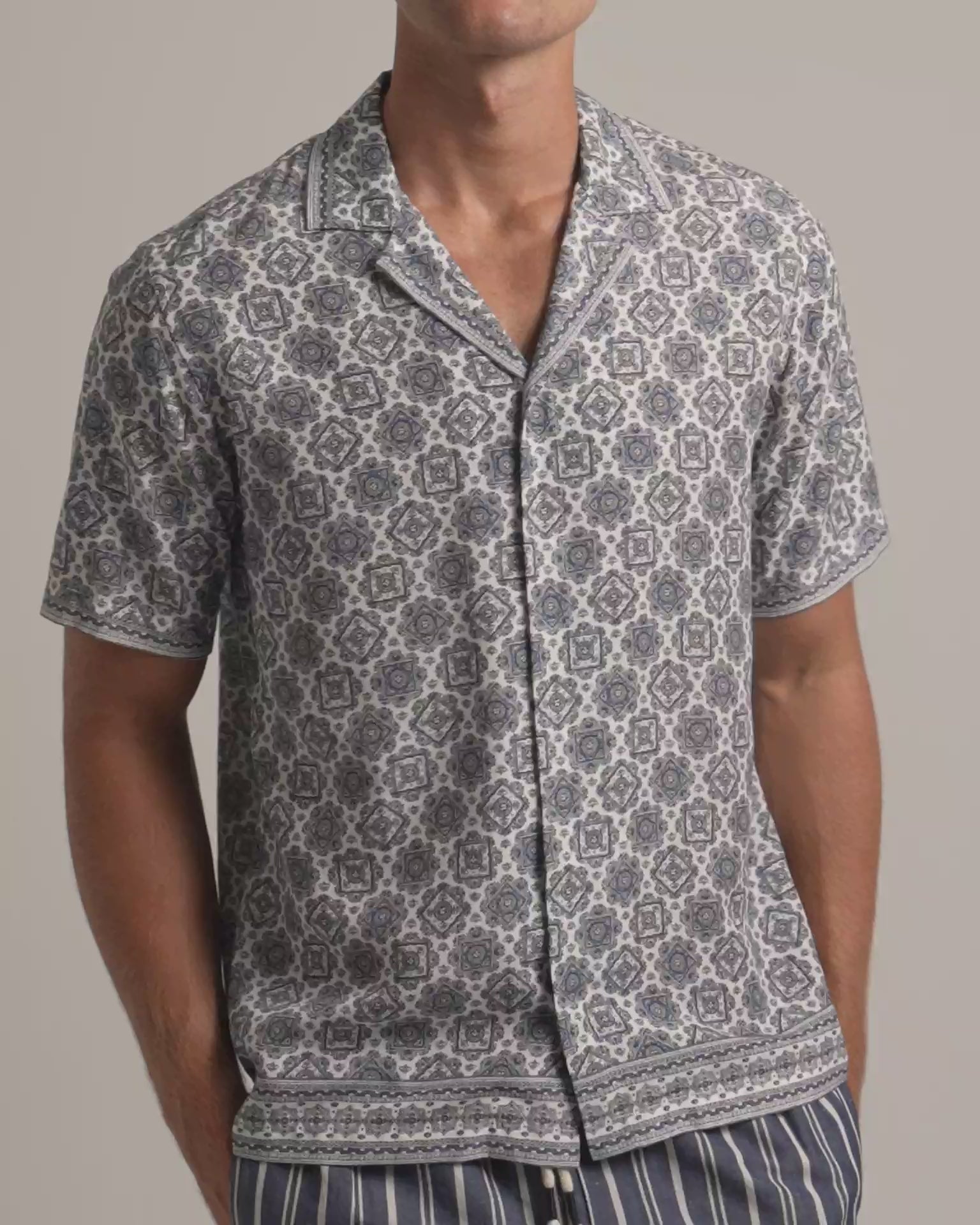 Man wearing white and grey geometric printed shirt with striped trousers against a plain background
