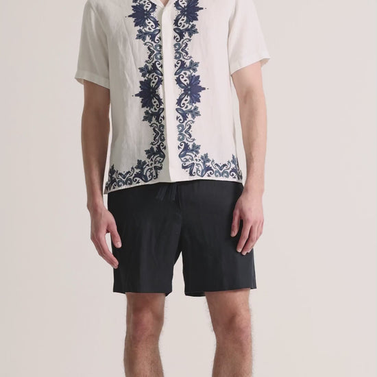 Man wearing printed white linen shirt with navy shorts on a plain background