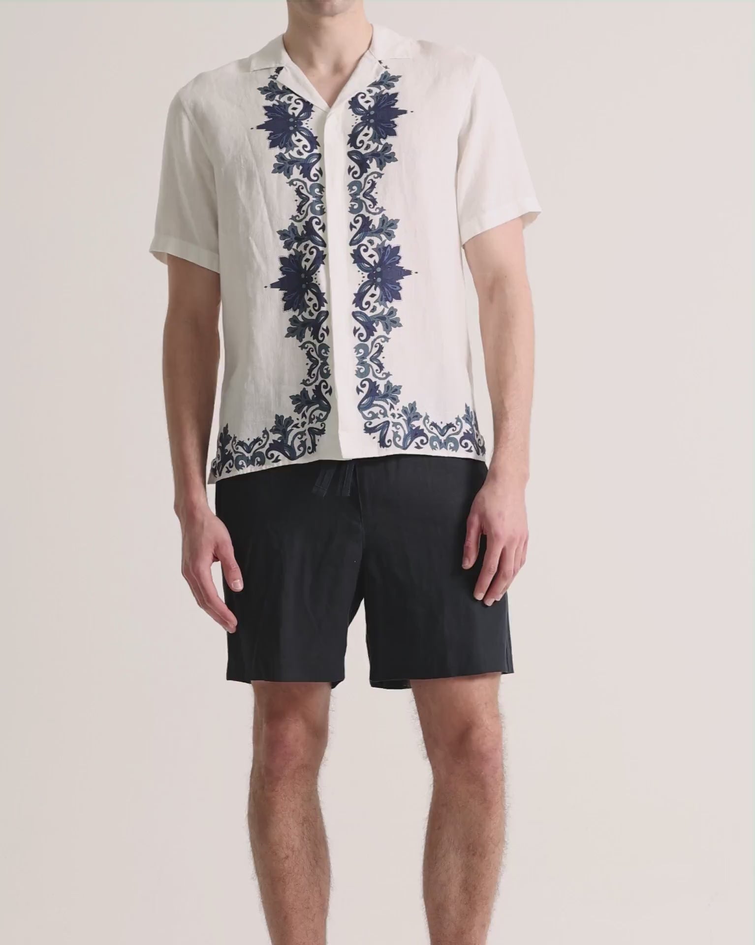 Man wearing printed white linen shirt with navy shorts on a plain background