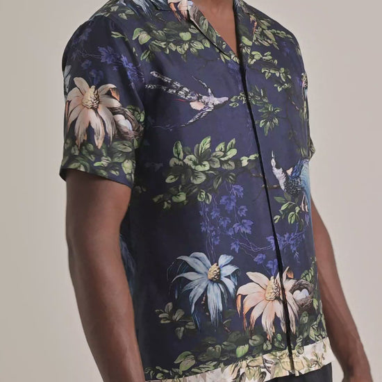 Man wearing navy shirt with botanical print in green, blue and pink. Modelling with dark brown trousers against a plain backdrop