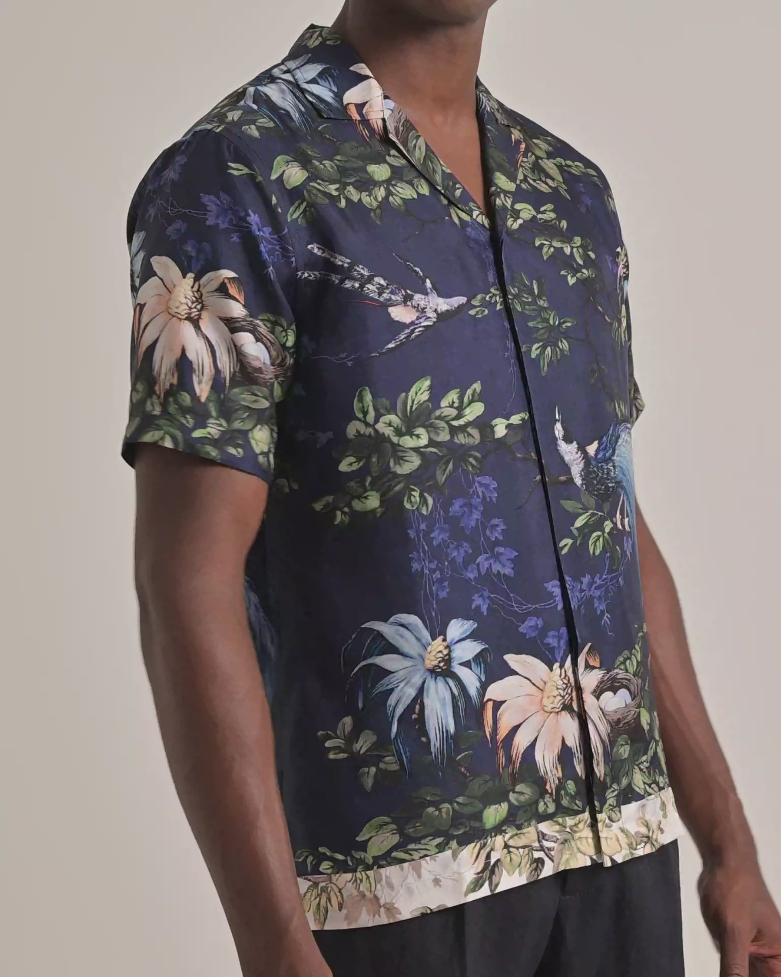 Man wearing navy shirt with botanical print in green, blue and pink. Modelling with dark brown trousers against a plain backdrop
