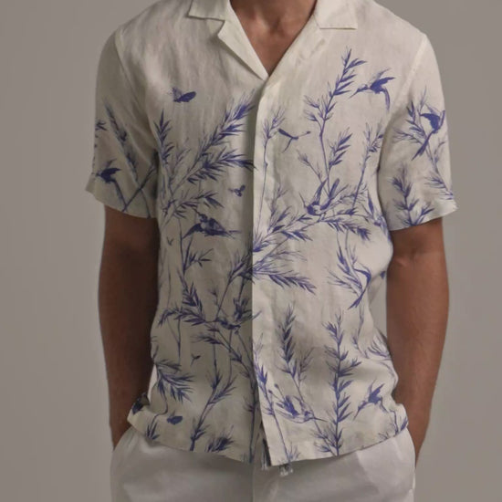 Man wearing white linen shirt with blue botanical print and white trousers on a plain background