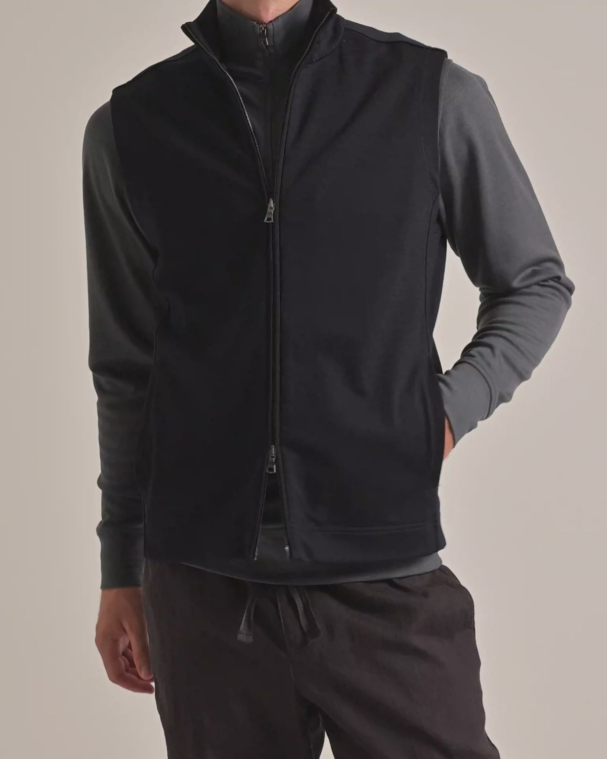 Man wearing lightweight gilet with grey quarter-zip sweatshirt underneath and dark brown trousers on a plain background
