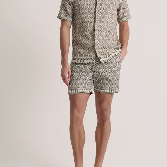 Man wearing beige printed shirt with shorts on a plain background