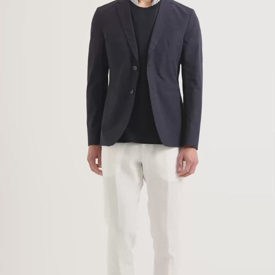 Man wearing navy blazer, navy jumper and stripe shirt underneath and white trousers