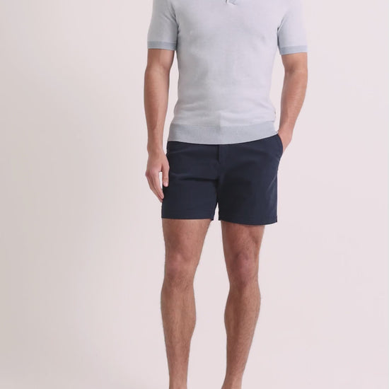 Man wearing blue knitted polo shirt with navy shorts on a plain background