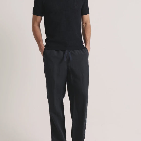 Man wearing navy knitted polo shirt with linen trousers on a plain background
