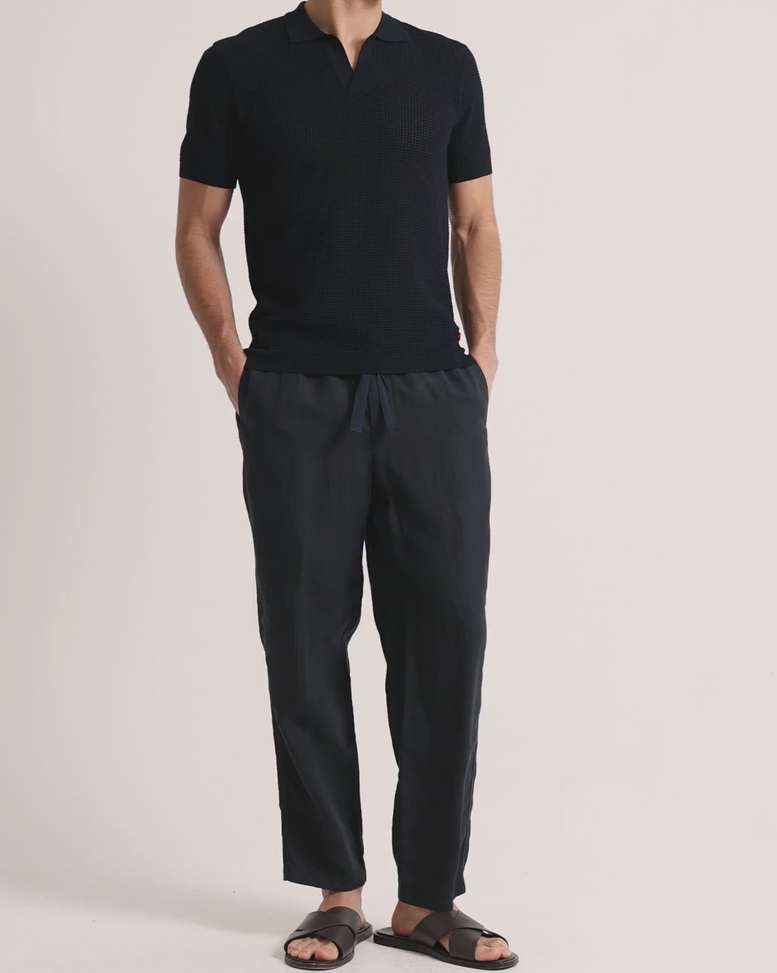 Man wearing navy knitted polo shirt with linen trousers on a plain background