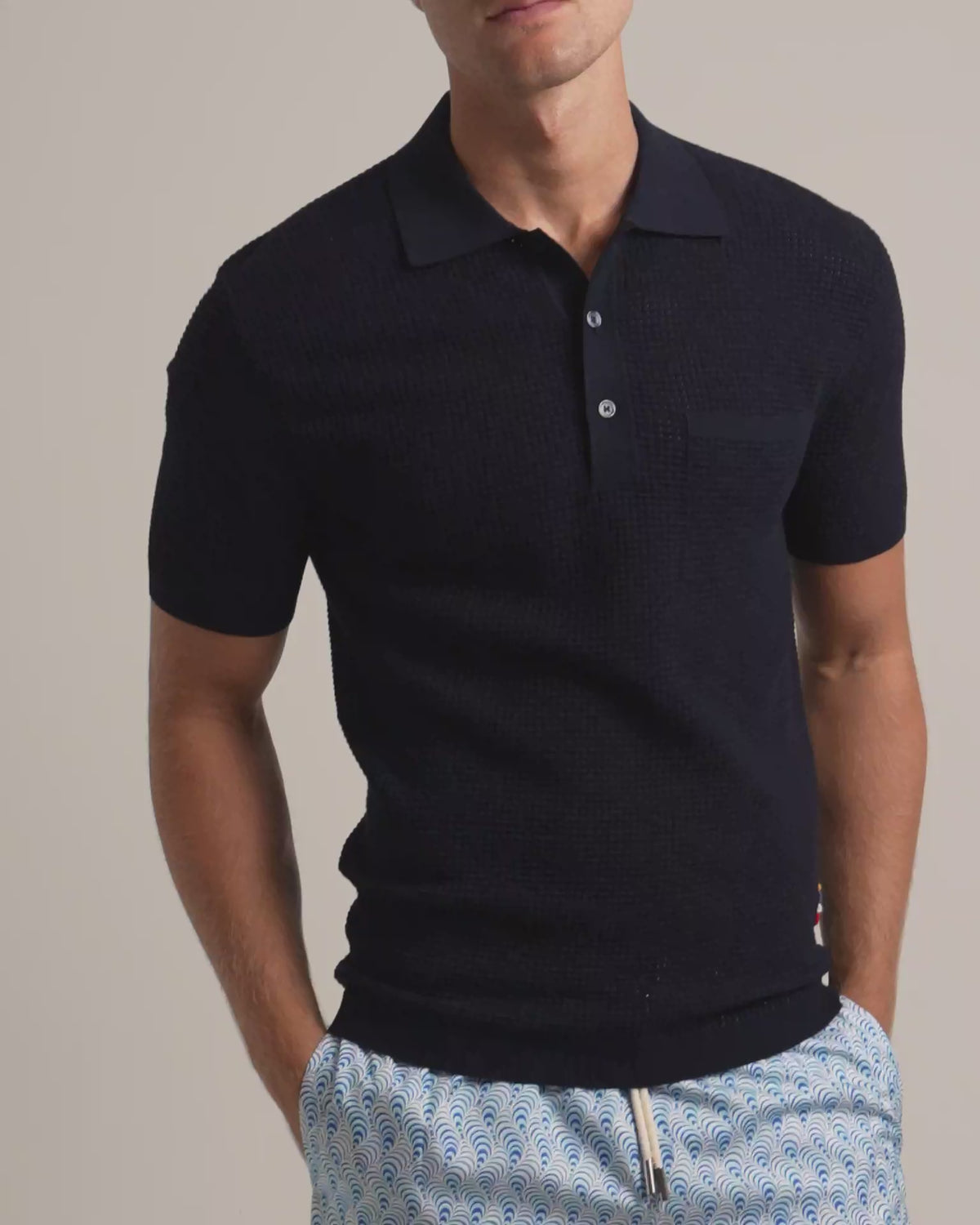 Man wearing navy mesh stitch polo shirt with chest pocket and blue patterned swim shorts against a plain background