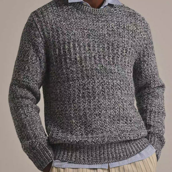 Man wearing grey knitted jumper in grey and white mix crochet. Worn with a blue shirt underneath and beige striped trousers against a plain background.