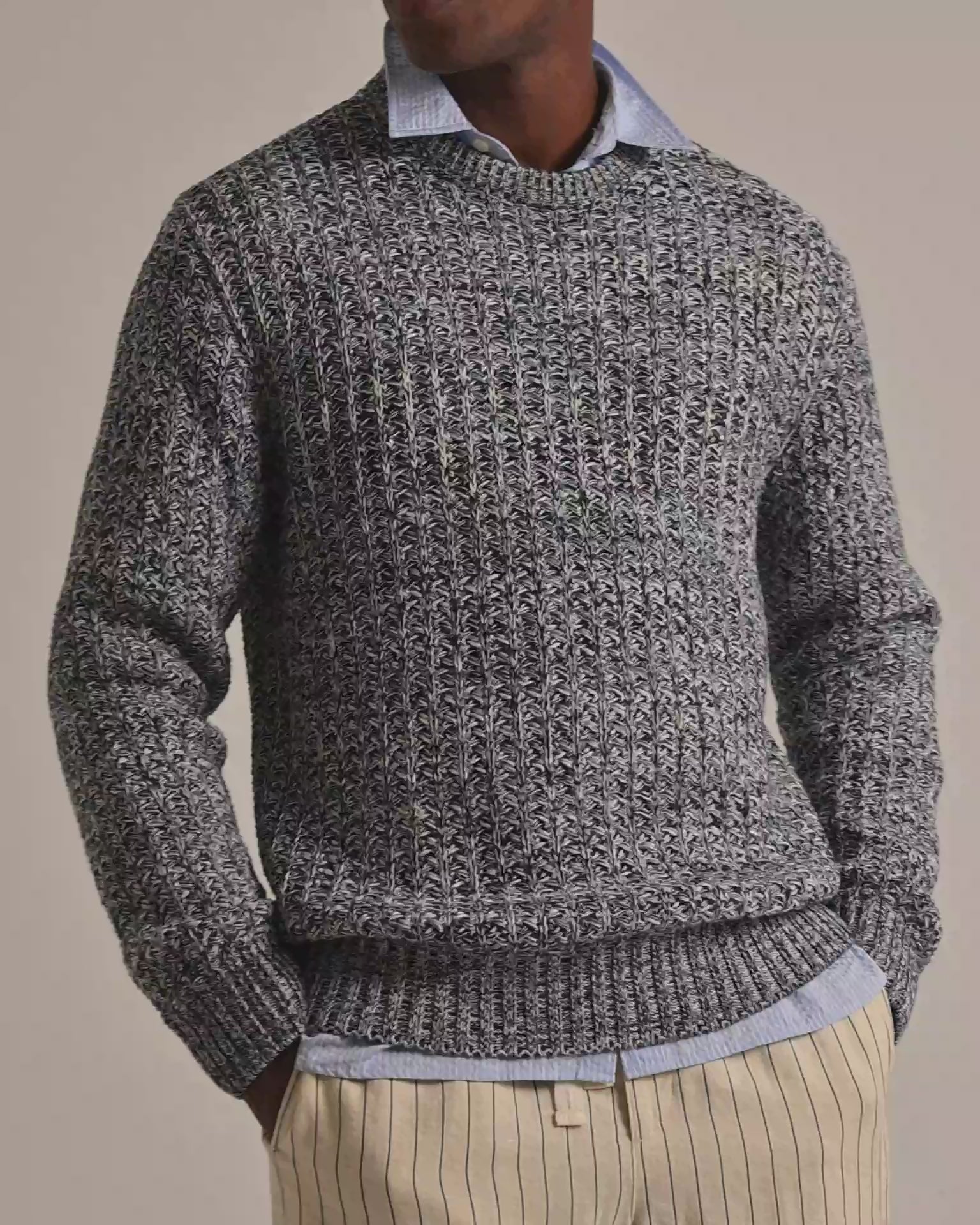 Man wearing grey knitted jumper in grey and white mix crochet. Worn with a blue shirt underneath and beige striped trousers against a plain background.