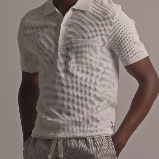 Man wearing white mesh stitch polo shirt with chest pocket and fine striped trousers against a plain background