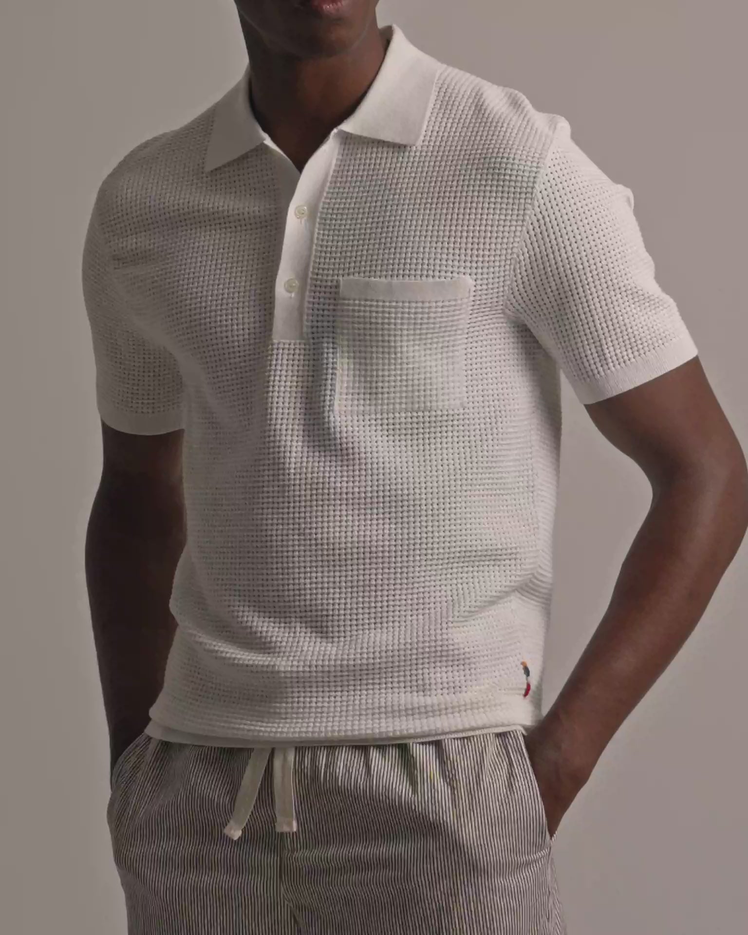 Man wearing white mesh stitch polo shirt with chest pocket and fine striped trousers against a plain background