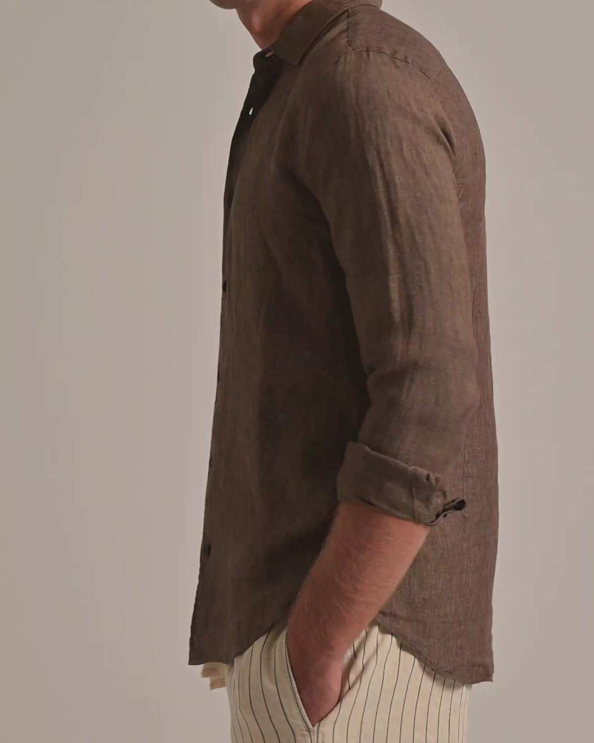 Man wearing brown linen shirt with light beige striped trousers on a plain background