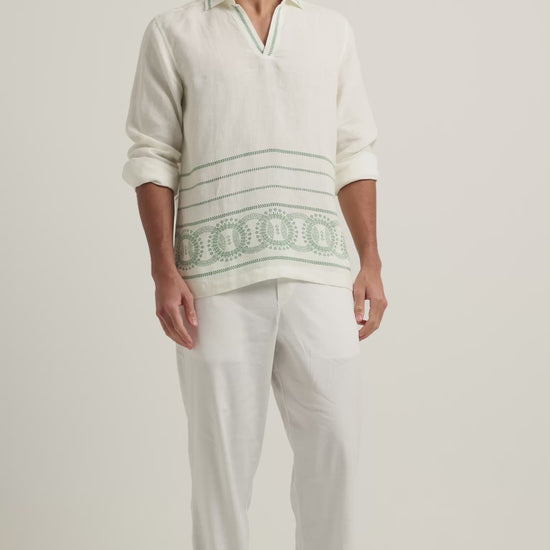 Man wearing long sleeve white shirt with green embroidery and white trousers on a plain background