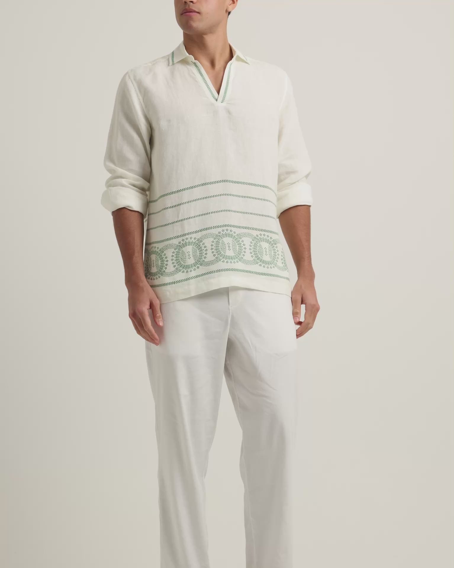Man wearing long sleeve white shirt with green embroidery and white trousers on a plain background