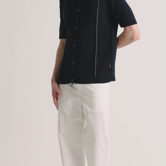 Man wearing navy knitted shirt with white trousers on a plain background