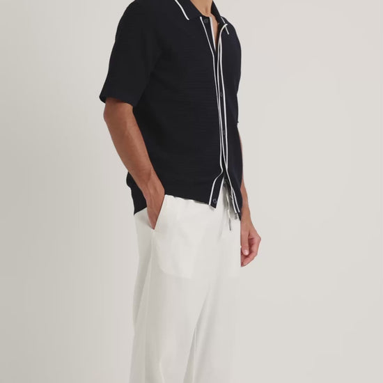 Man wearing black textured knitted black shirt with white trim detailing and white drawstring trousers on a plain background