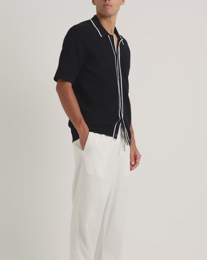 Man wearing black textured knitted black shirt with white trim detailing and white drawstring trousers on a plain background