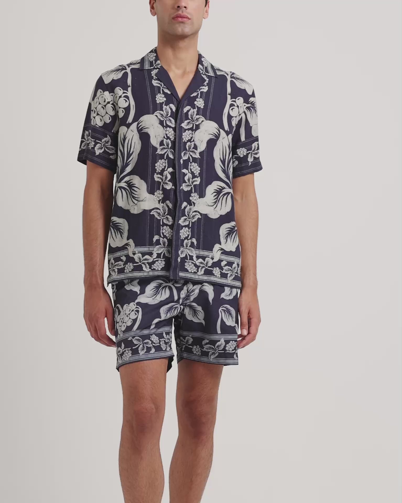 Man wearing floral print navy and white shirt with matching swim shorts on a plain background